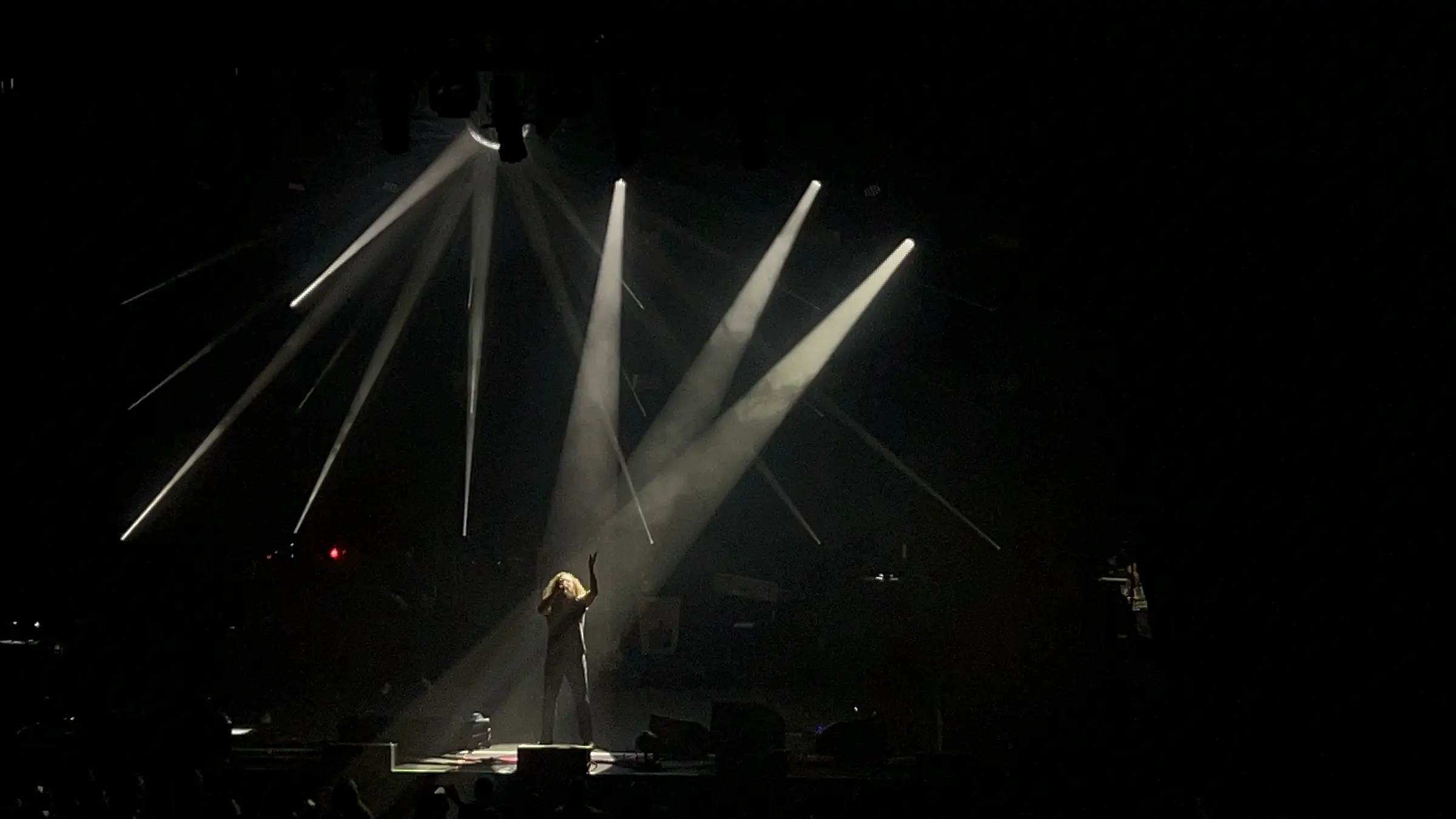 A performer on a dark stage at the lower centre, one arm raised, under fanning white spotlight beams cutting through haze